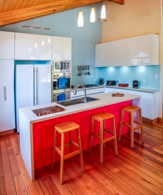 A modern kitchen featuring a central island with a colorful red base and three wooden stools. The kitchen is equipped with stainless steel appliances, including a refrigerator and oven. Bright blue walls complement the light wood ceiling and polished wooden floor, enhancing the spacious feel.
