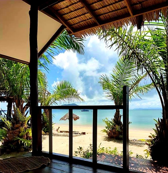 The image shows a view from a shaded deck, featuring palm trees framing a serene beachfront. Soft clouds drift in a bright sky above tranquil waters, which reflect the light. Beach chairs and a sunshade are positioned on the sandy shore, inviting relaxation.