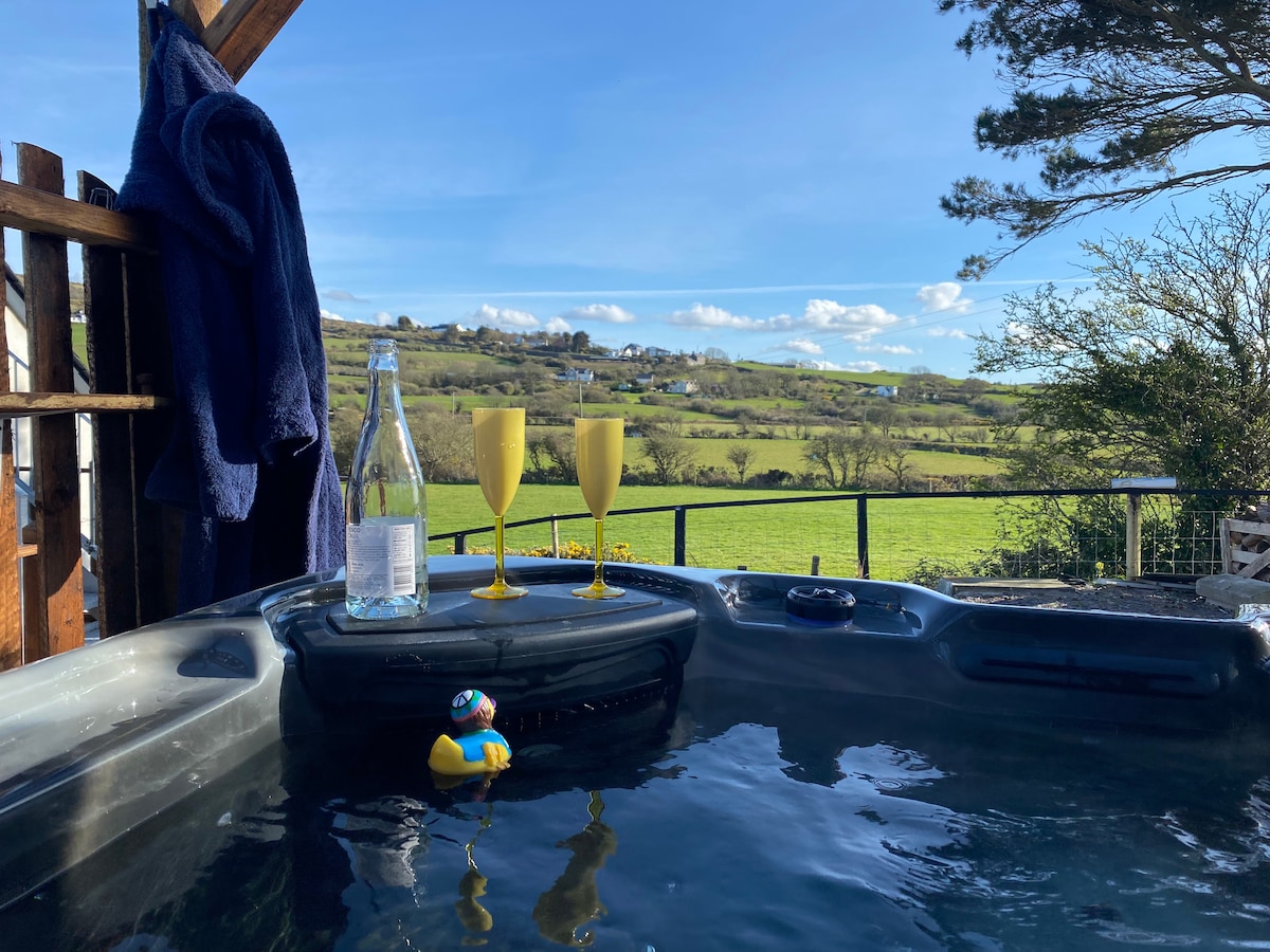 A hot tub filled with clear water is positioned on a private deck, overlooking expansive green hills under a bright blue sky. Two yellow glasses and a bottle of water rest on the edge, while a small rubber duck floats nearby.