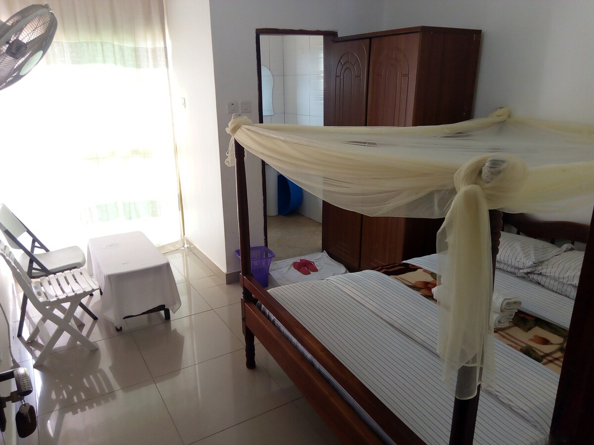 A well-furnished room features a wooden four-poster bed draped with a mosquito net. A large mirror reflects the space, which includes a folding chair and a small table. Natural light enters through a partially covered window, enhancing the room's bright and airy feel.