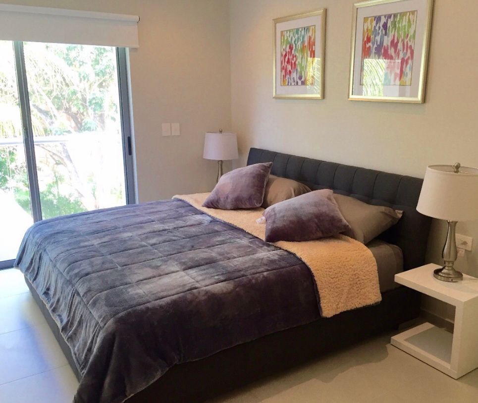 A serene bedroom is presented, featuring a spacious bed with a gray blanket and soft accent pillows. Two framed artworks adorn the wall above the bed, and a bedside lamp sits on a small table. A large window allows natural light to fill the room.