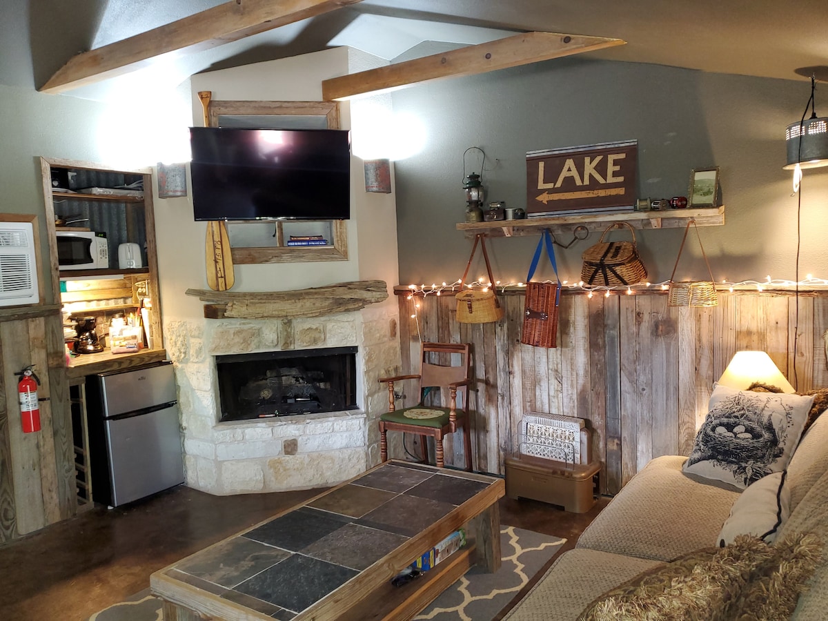 A cozy living area features a mounted television above a stone fireplace, with a rustic coffee table in the center. Shelves display decorative items and twinkling string lights add a warm touch. A sofa is positioned invitingly nearby, alongside a mini fridge and coffee bar area.
