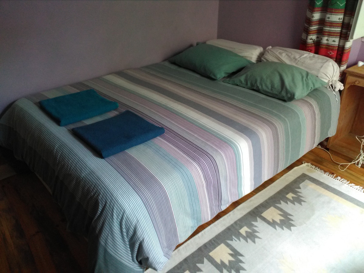 A spacious bed dressed with a striped blanket and accompanied by two folded towels is positioned in the room. The walls are painted in a soft purple hue, and colorful curtains frame a window, allowing natural light to enhance the space.