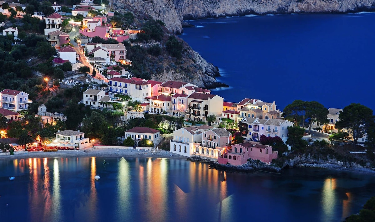 A panoramic view of a coastal village is captured at dusk, showcasing homes with warm lights reflecting on the calm waters below. The surrounding hills and dark blue sea create a serene backdrop, while the charming architecture emphasizes the village's tranquil atmosphere.