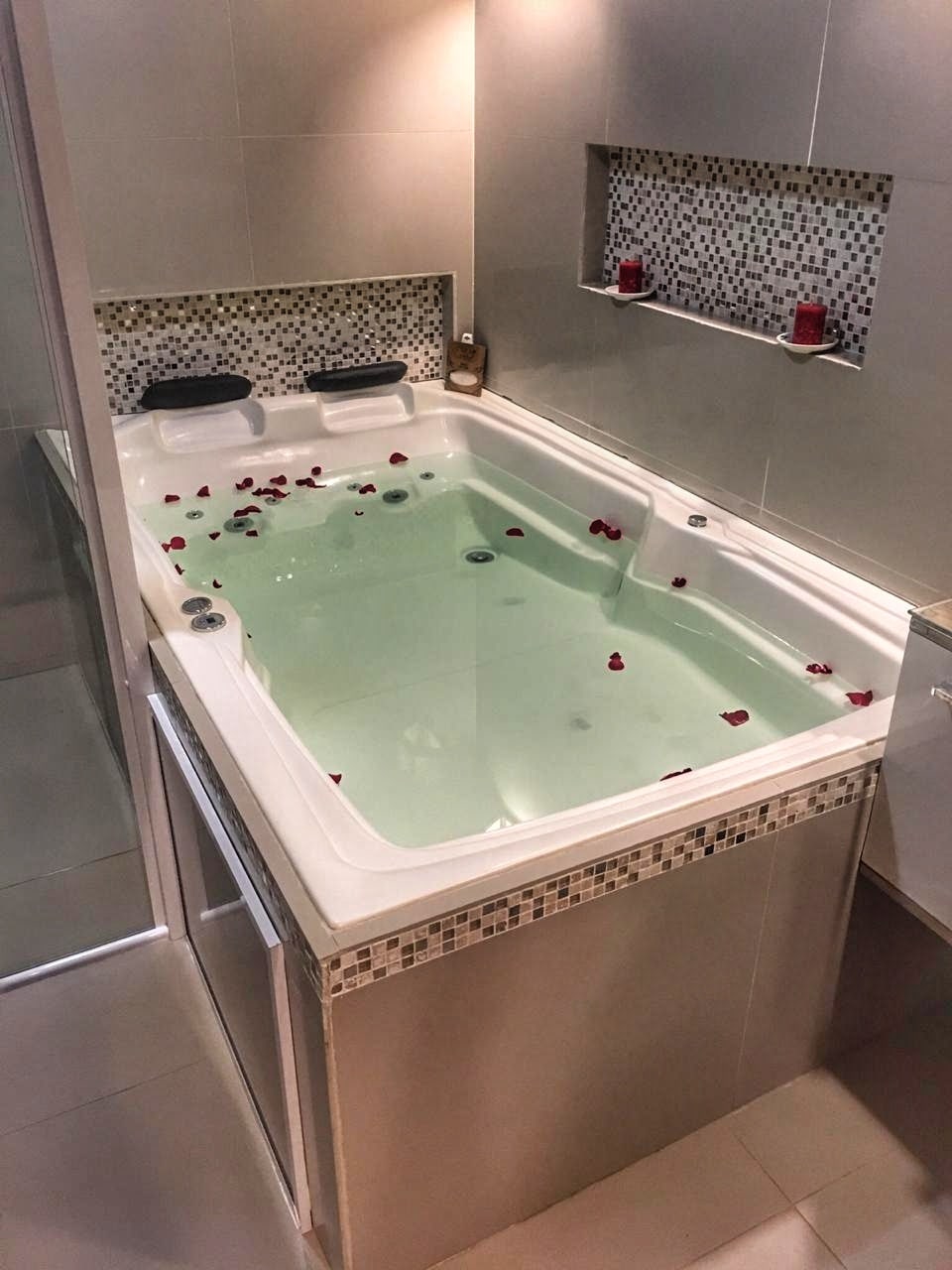 A private suite is equipped with a spacious whirlpool tub, adorned with rose petals on the surface of the water. Soft lighting highlights the surrounding tiles and decorative accents, creating a calming and relaxing atmosphere within the bathroom space.