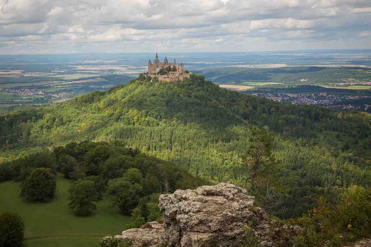 A panoramic view of a castle perched atop a hill, surrounded by lush green forests and rolling landscapes under a cloudy sky. The distant horizon stretches out, revealing fields and valleys, creating a serene and majestic atmosphere.