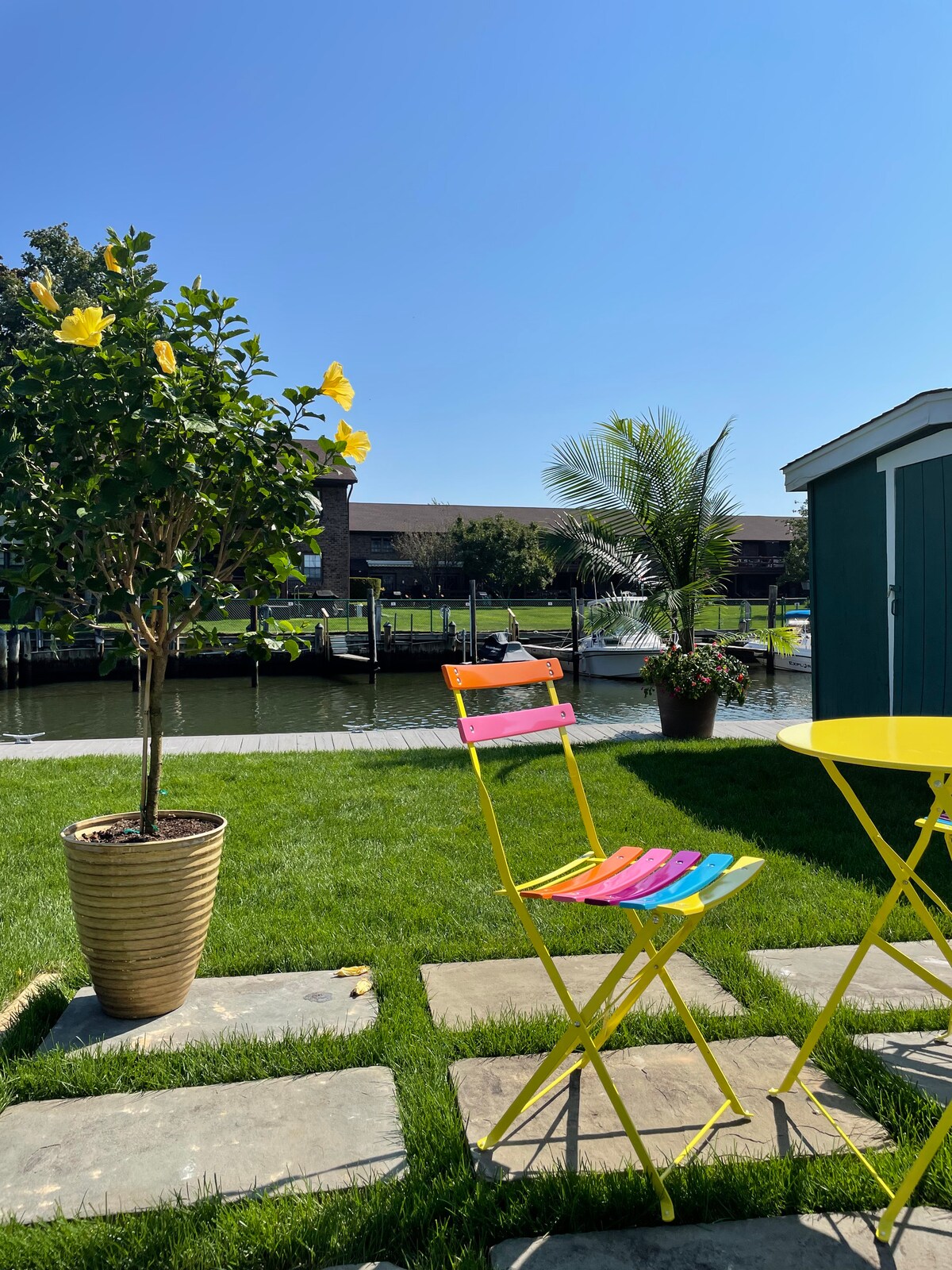 A vibrant outdoor seating area is highlighted by a yellow bistro table and colorful chairs. Tropical plants, including a hibiscus tree, add greenery against a backdrop of blue skies and the calm water of the bay, enhancing the tranquil atmosphere.