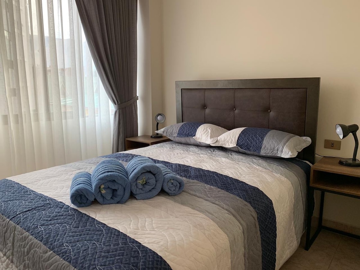 A cozy bedroom is featured with a spacious bed dressed in a blue and white quilt. Soft light filters through sheer curtains, enhancing the warm tones. Two bedside tables are placed at either side, each equipped with a reading lamp. Rolled towels are neatly arranged at the foot of the bed.