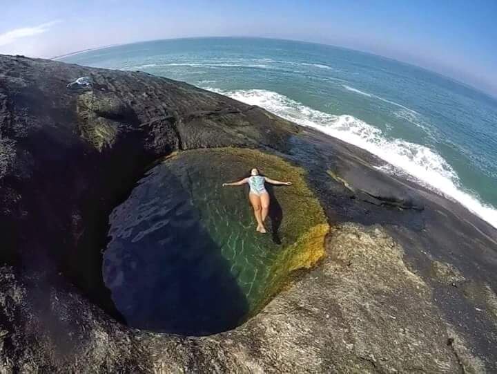 A natural rock pool is visible alongside the shoreline, surrounded by rugged dark stones. Crystal-clear water reflects the blue sky and waves crashing nearby, creating a serene and inviting atmosphere for relaxation and enjoying the ocean's proximity.