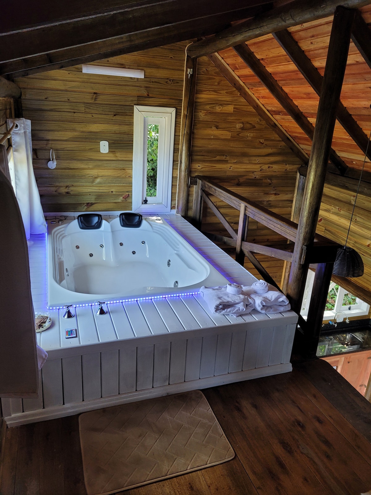 An indoor hot tub is positioned in a wooden loft area, surrounded by natural light from a small window. The hot tub features seating and is illuminated by soft LED lights along the edges. Wooden beams and warm tones enhance the rustic aesthetic of the space.