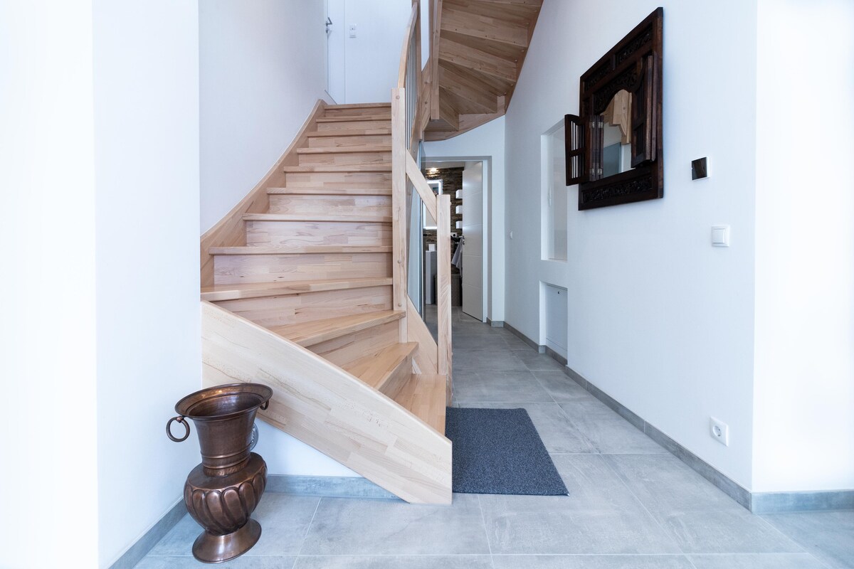 A wooden staircase with smooth oak steps is situated against a white wall, leading to the upper floor. A decorative metal vessel rests on the tiled floor, while a dark-framed mirror adds depth to the entrance space.