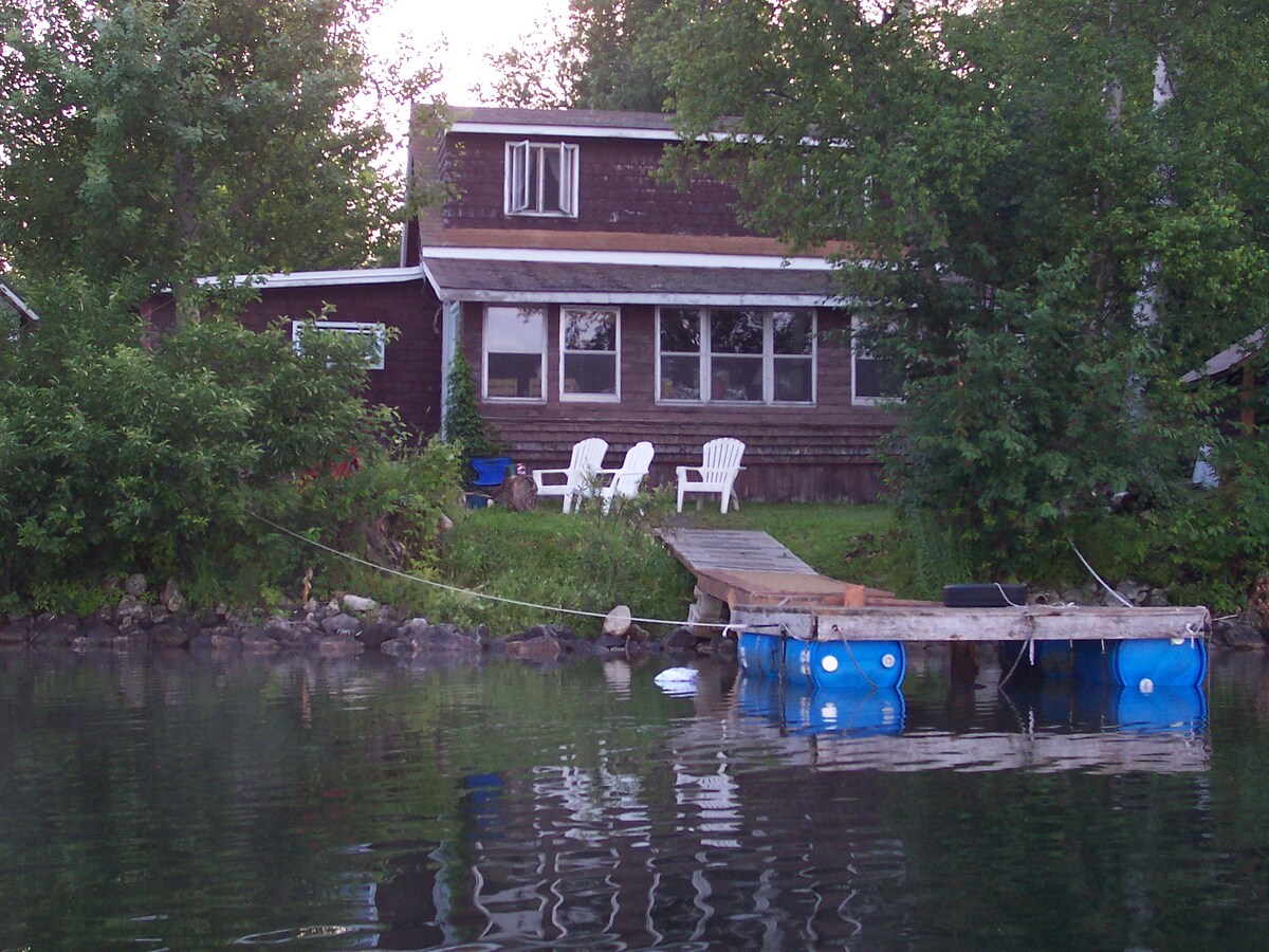 A rustic lakefront cottage is set amid greenery, featuring large windows and a welcoming porch. Two white Adirondack chairs are positioned on the front lawn, with a wooden dock extending into the calm water, creating access to the lake.