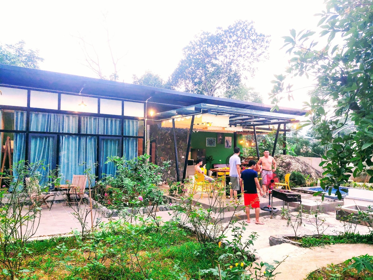 The modern house is surrounded by a lush garden, featuring a patio area with colorful chairs and a grill. Large glass doors enhance the connection with the outdoors, while guests engage in outdoor activities under a covered space, surrounded by greenery.
