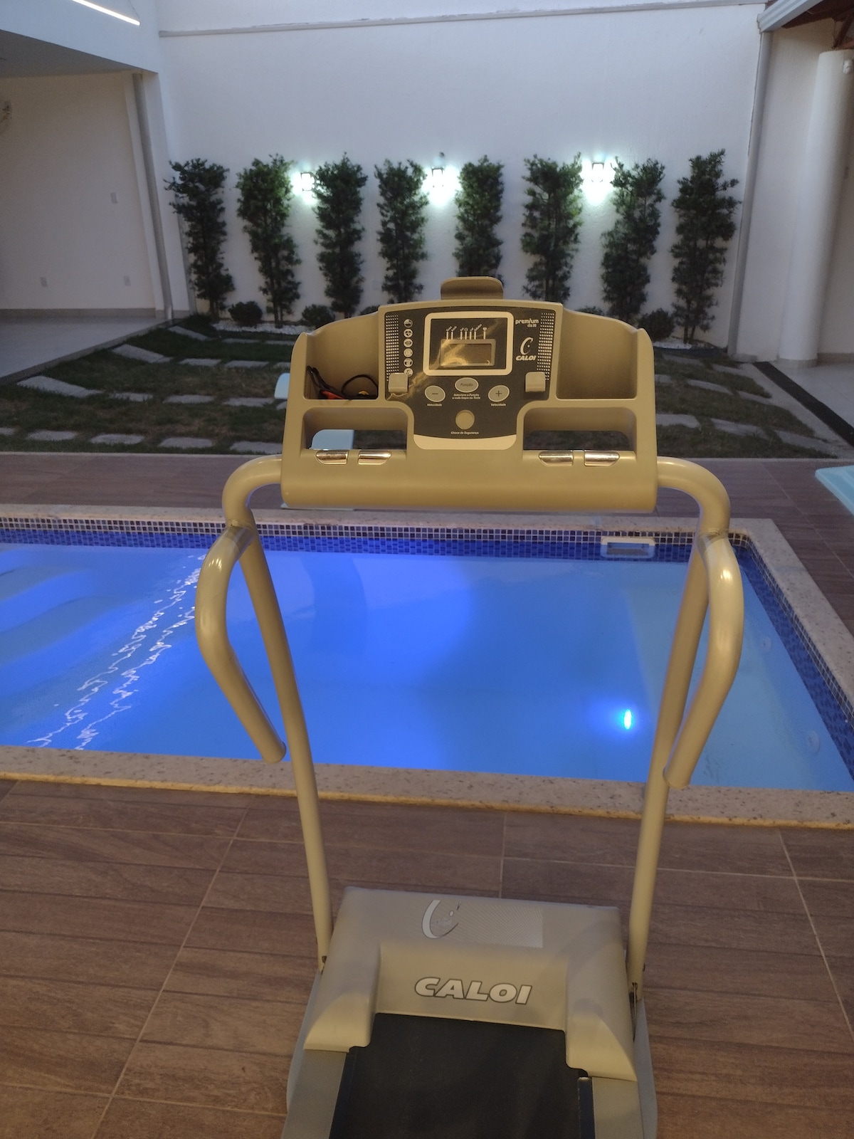 An electric treadmill is positioned in front of a sparkling pool, which is softly illuminated in blue. Surrounding foliage is visible, providing a serene outdoor backdrop with well-maintained grass and strategically placed lights.