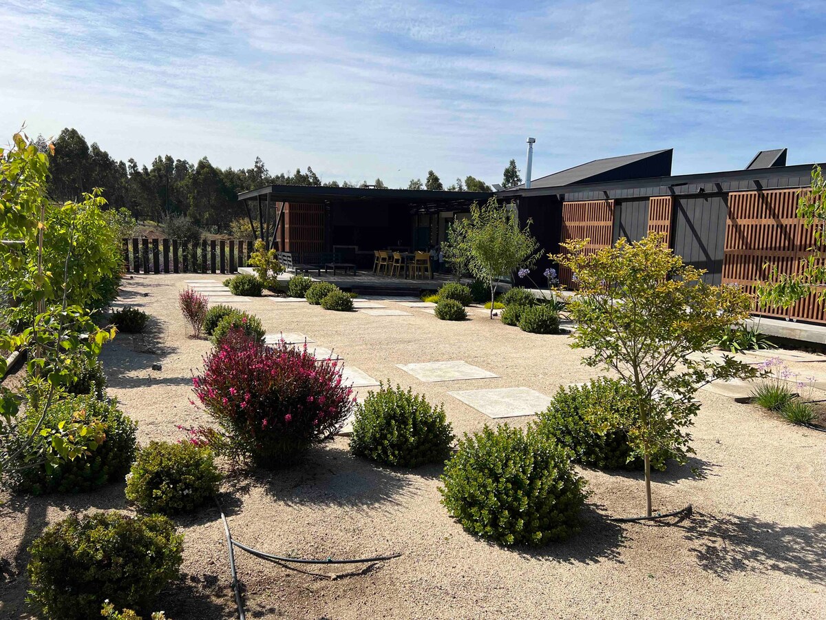 A well-maintained outdoor space features a variety of greenery, including small shrubs and flowering plants, framed by gravel pathways. The modern structure with wooden accents is visible in the background, complemented by a deck area with seating.