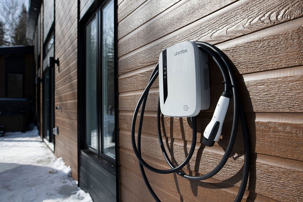 An electric vehicle charging station is mounted on the wooden exterior of the cottage. The charging cable is neatly coiled and hangs along the wall, with a snow-covered ground below. The surrounding area features large windows that provide a view of the exterior landscape.
