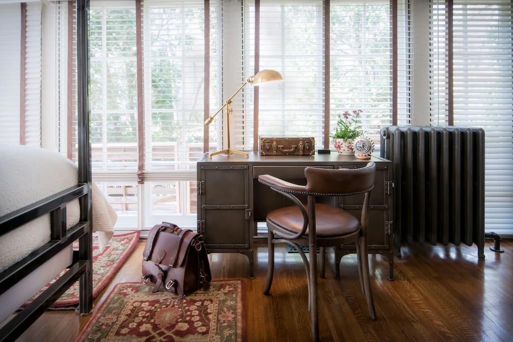 A sun-filled first floor bedroom is accented with a sleek steel framed canopy bed, handsome desk, and pair of sturdy leather library chairs by the fireplace.