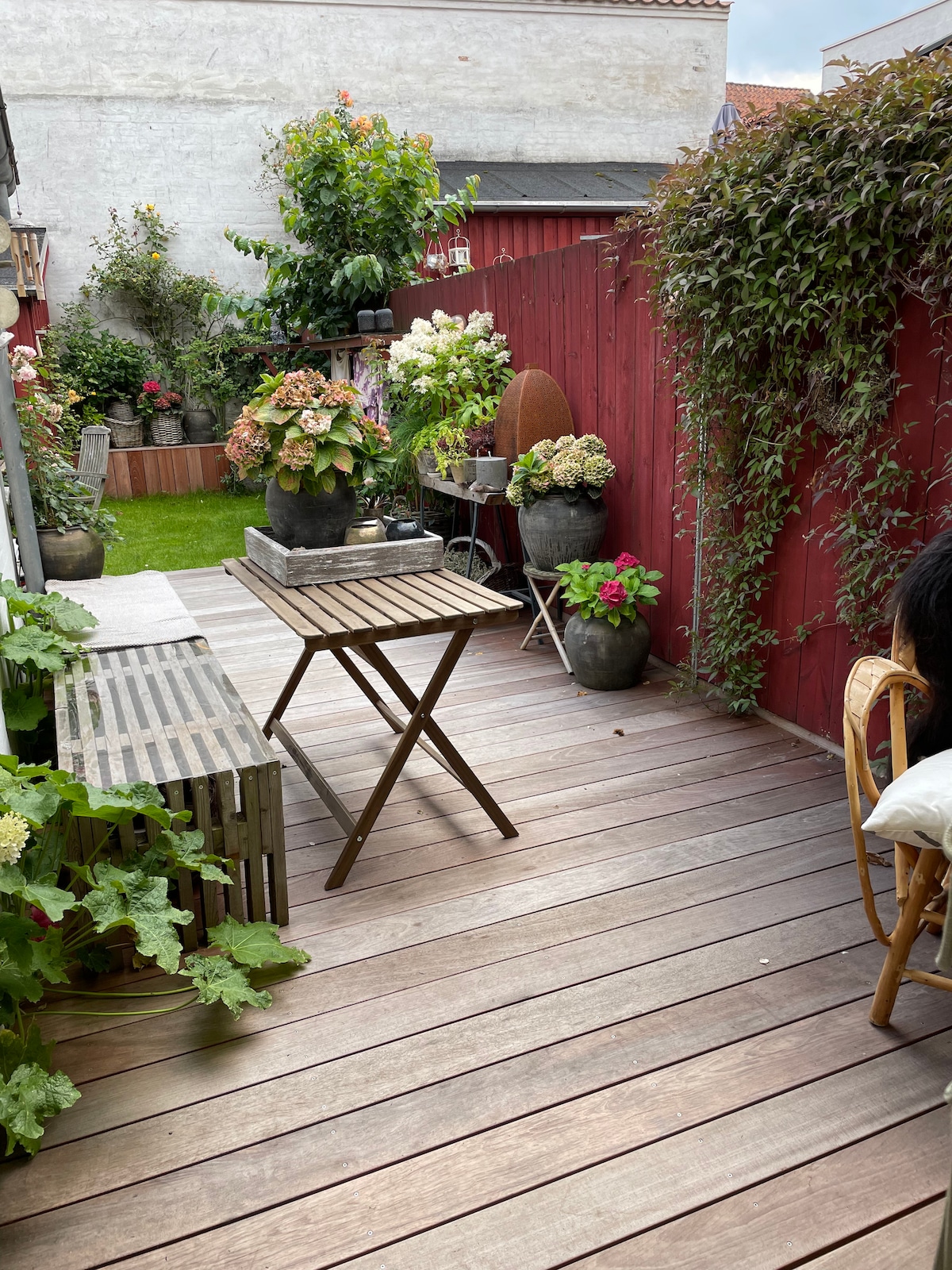 A charming private courtyard is displayed, featuring wooden decking and various potted plants. A small table is positioned centrally, surrounded by greenery and colorful flower arrangements. The backdrop includes a red fence and additional foliage, creating a tranquil outdoor space.