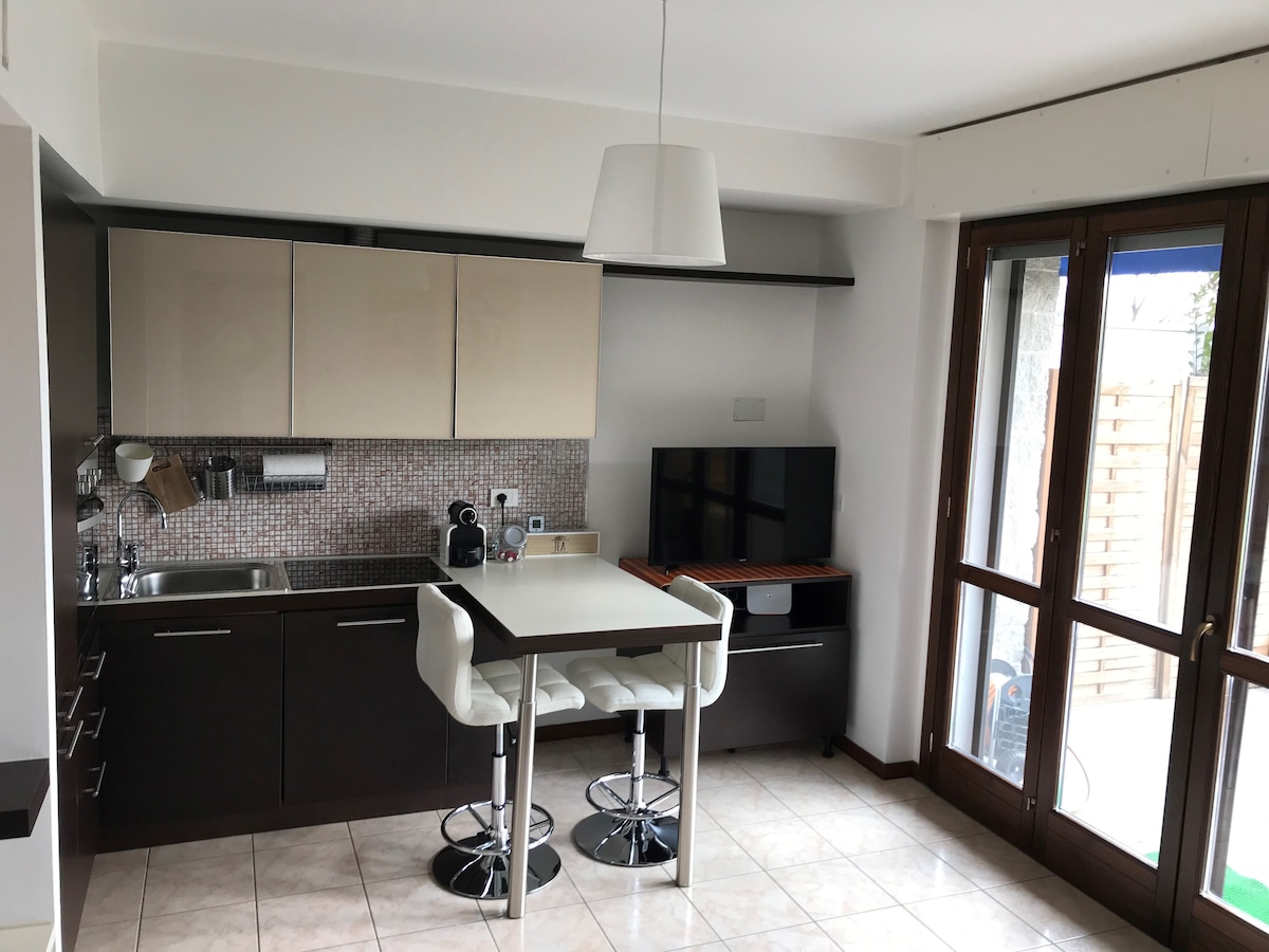 A modern kitchen area features sleek cabinetry and a minimalist design. A small dining table is accompanied by two stools. A television is positioned on a stand beside the window, which opens to the private garden. Natural light brightens the space.