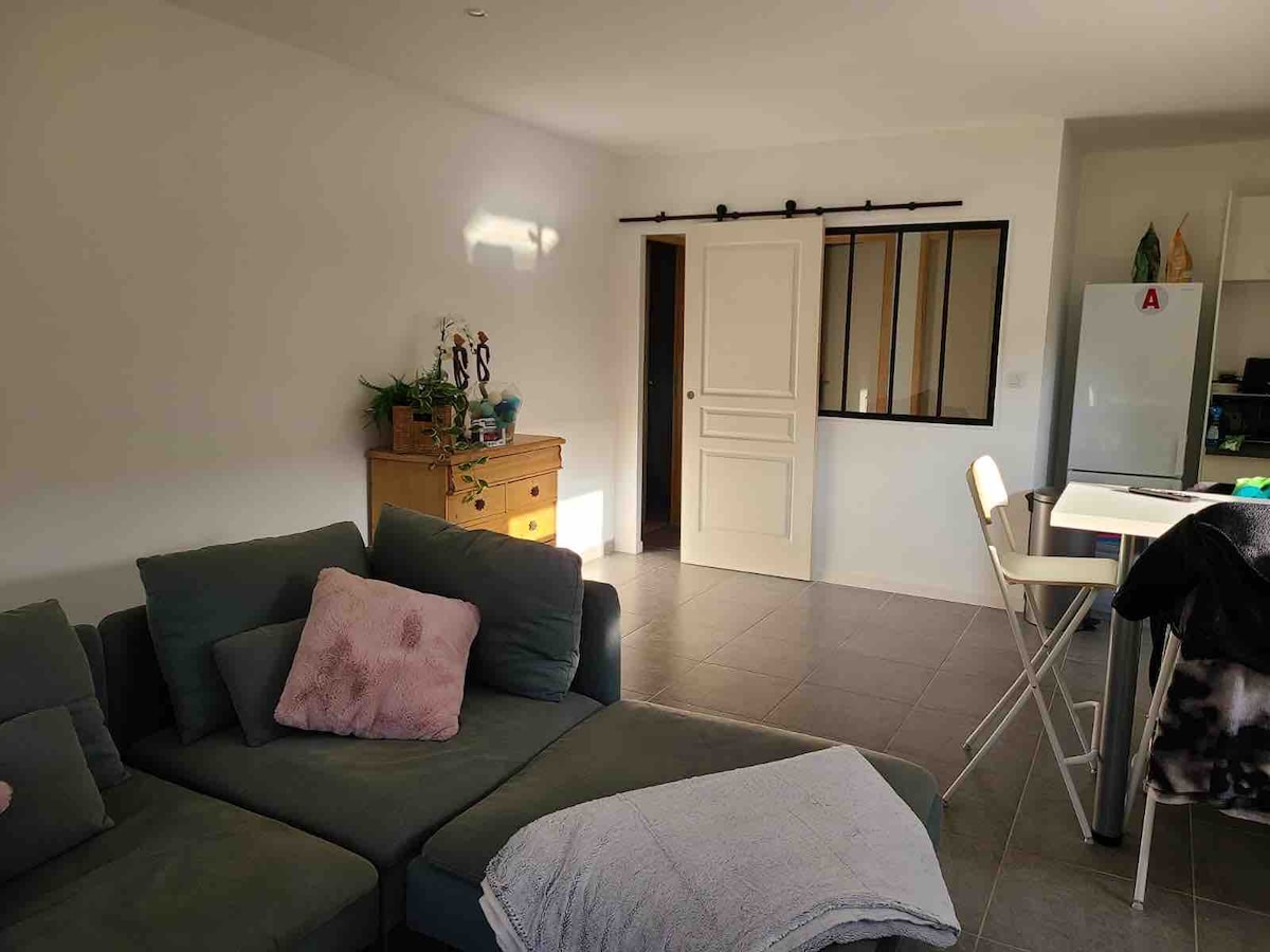 A spacious living area is visible, featuring a comfortable gray sofa adorned with a soft pink pillow. A wooden dresser adds warmth to the room, while a modern kitchen with white cabinetry can be seen in the background. Natural light enters through the window, enhancing the open space.