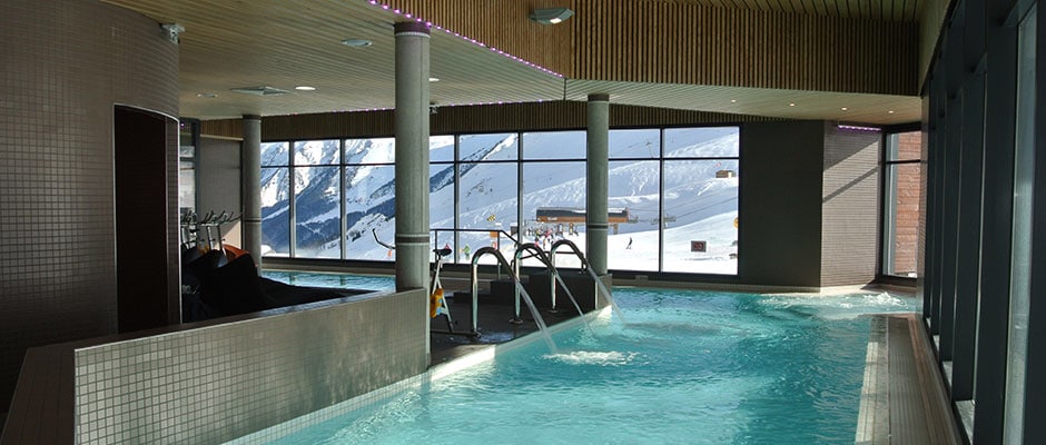 An indoor pool area is depicted, featuring large windows that offer views of the snow-covered mountains outside. The inviting turquoise water is complemented by modern architectural elements, including wooden ceilings and tiled surfaces, creating a bright and spacious environment.