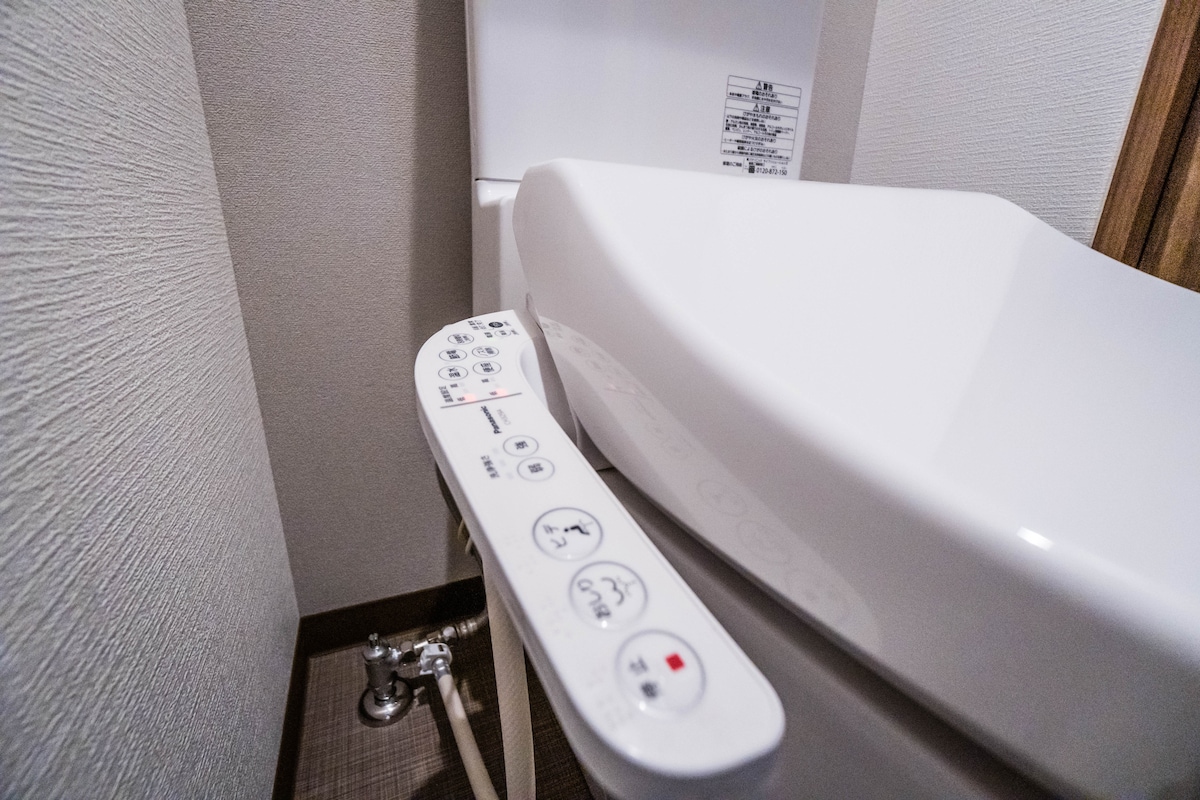 The image displays a modern toilet equipped with a control panel on the side. The panel features various buttons for adjusting settings, emphasizing convenience and advanced functionality. The surrounding walls have a neutral texture, and the floor is designed with a subtle pattern.