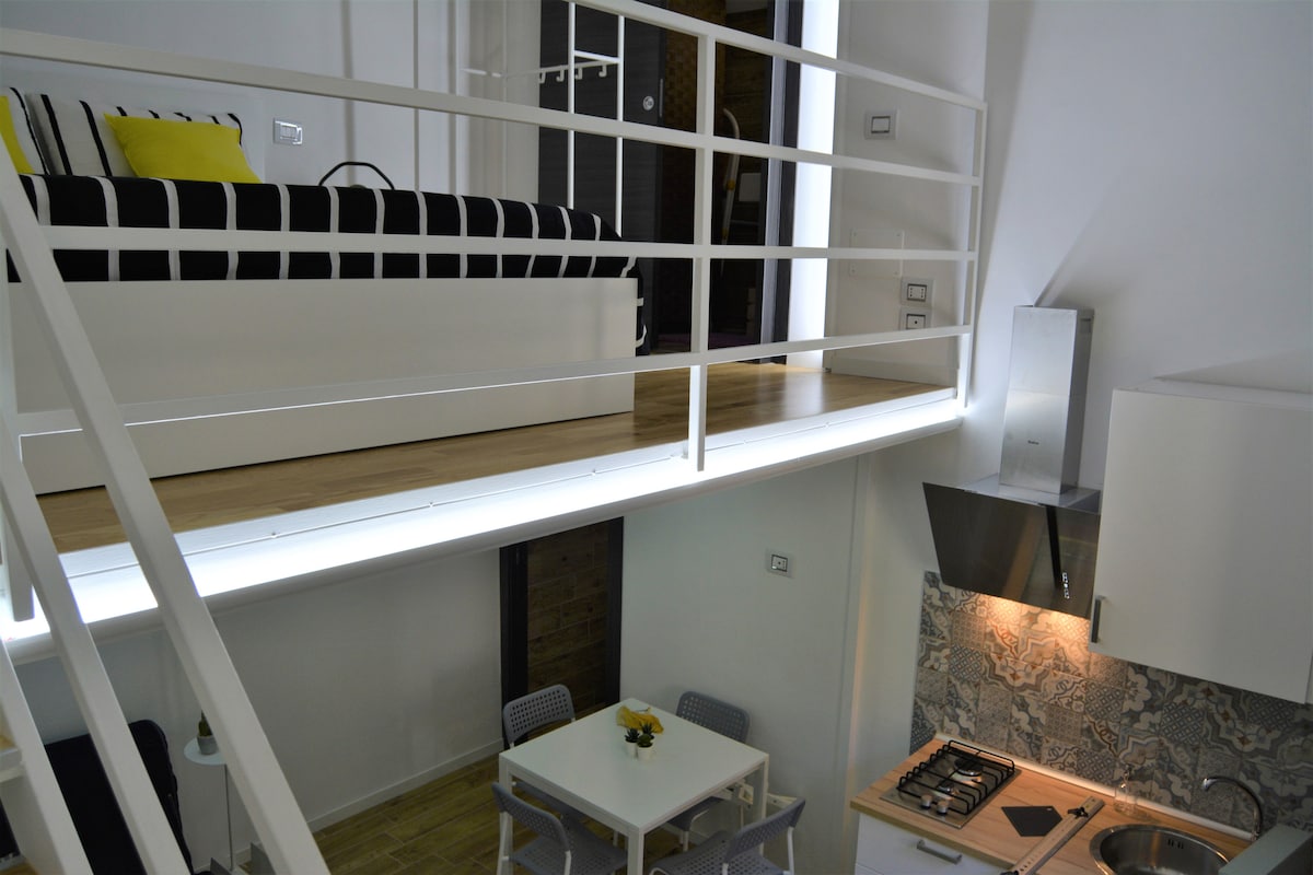 The interior of the loft showcases a modern design with a lofted sleeping area above a cozy kitchen and dining space. Soft lighting enhances the wooden floors, while the black and white striped couch adds a touch of elegance. A small table sits beside the kitchen, ready for dining.