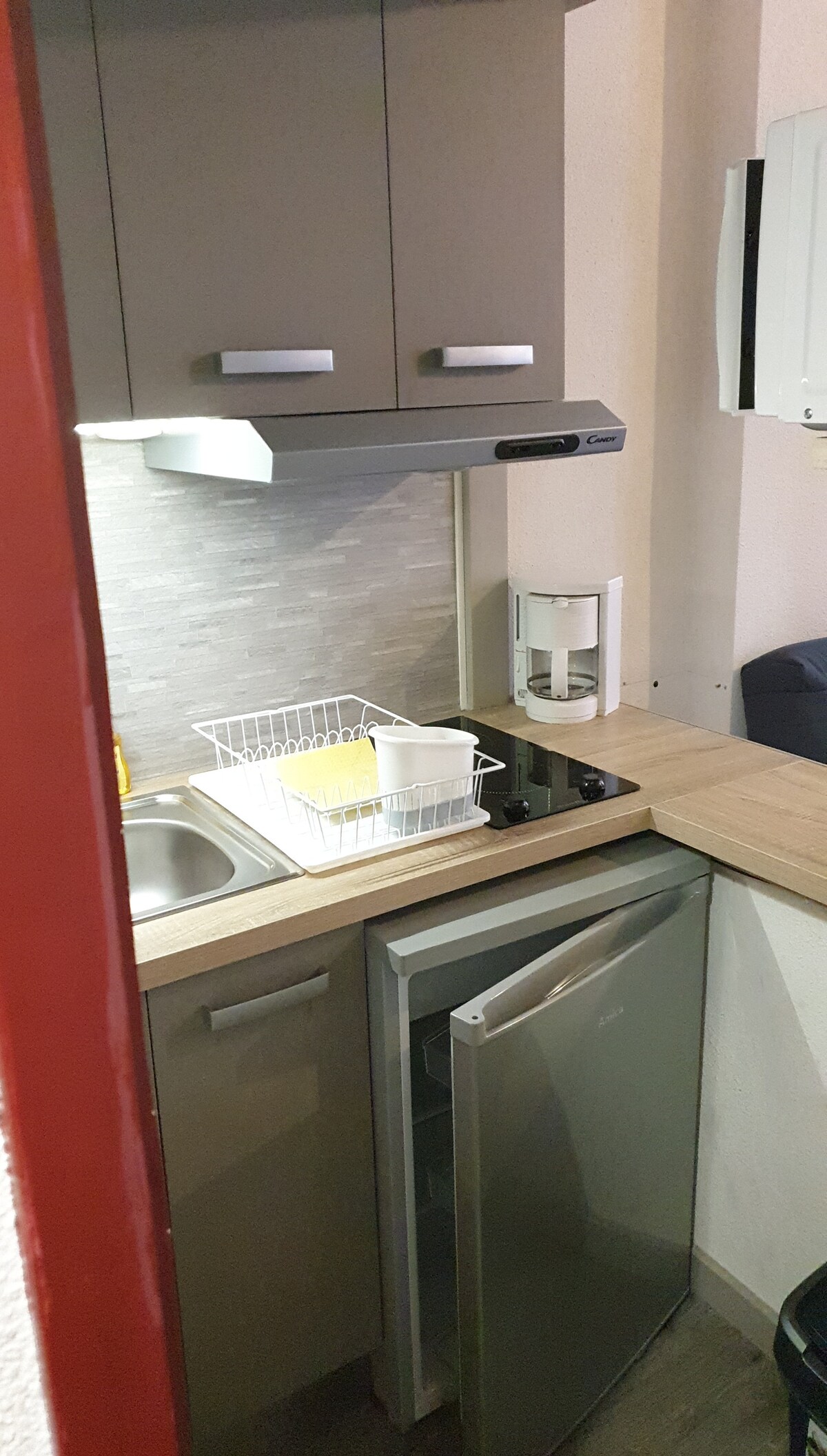 A compact kitchen space is equipped with modern cabinetry and essential appliances. A sink and a small refrigerator are present, accompanied by a two-burner cooktop. A dish rack holds dishes on the countertop, with soft lighting illuminating the area.
