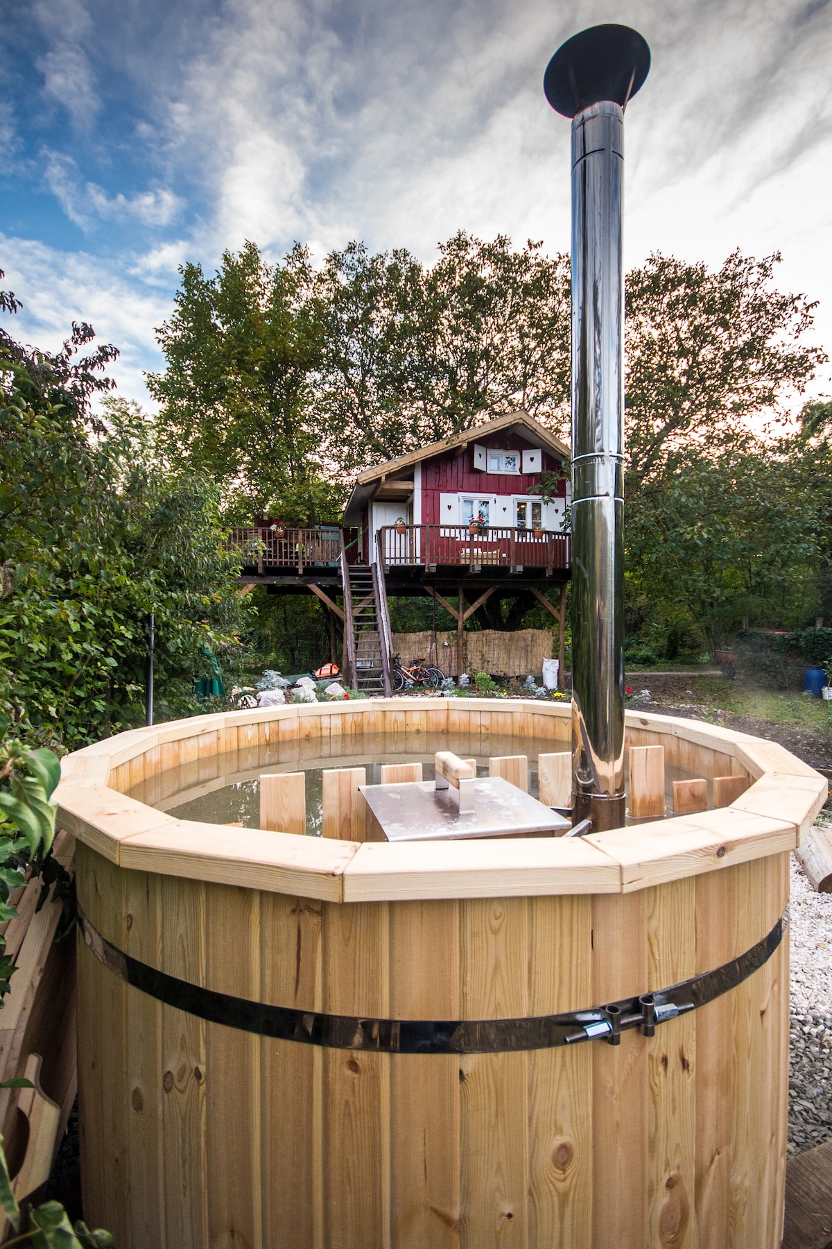 A wooden hot tub is situated in a garden, featuring a metal chimney for wood heating. In the background, a distinctive house with a sloped roof and deck can be seen, framed by lush greenery and a cloudy sky.