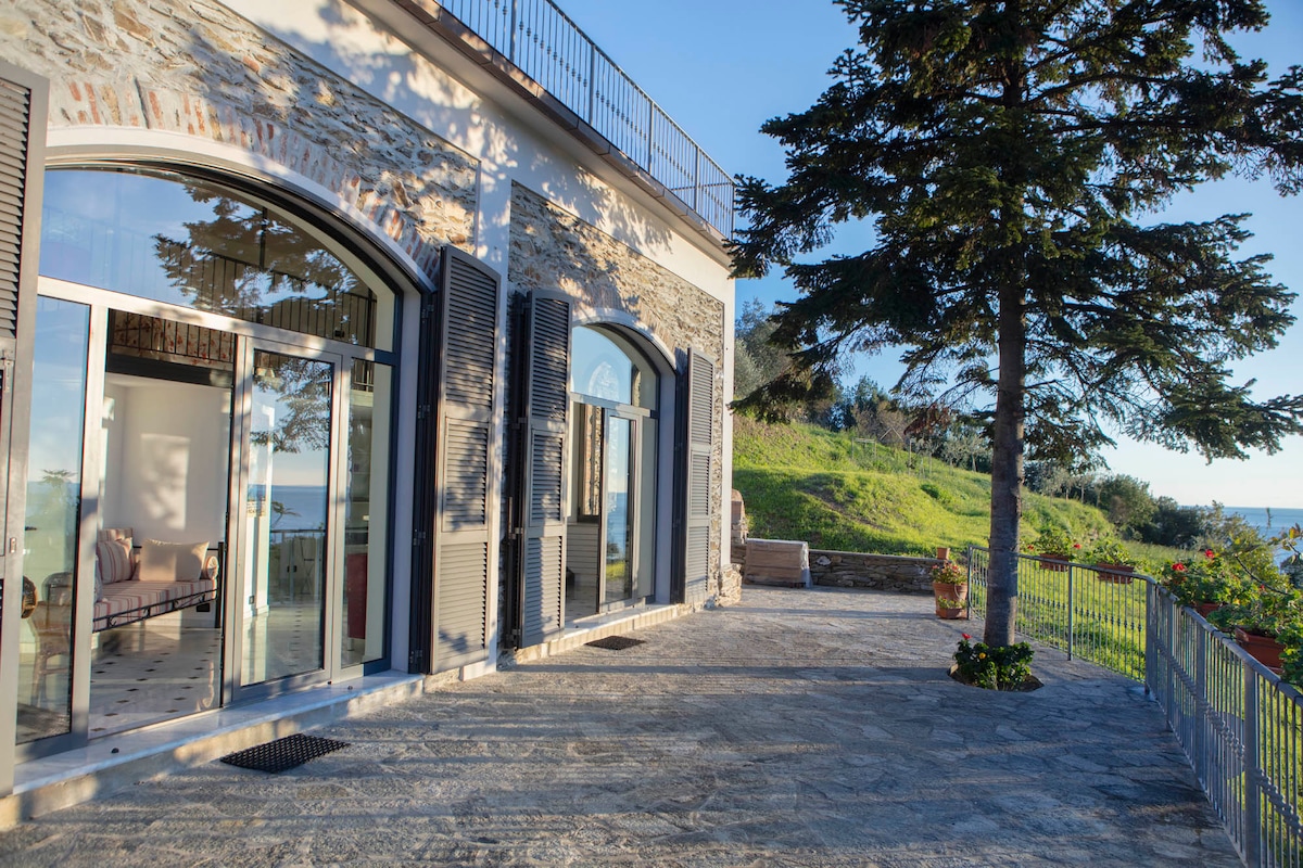 A spacious terrace is shown, featuring large glass doors that open to the interior. The area is surrounded by lush greenery and offers views of the sea. Sunlight casts a warm glow on the stone flooring, highlighting the inviting outdoor space.