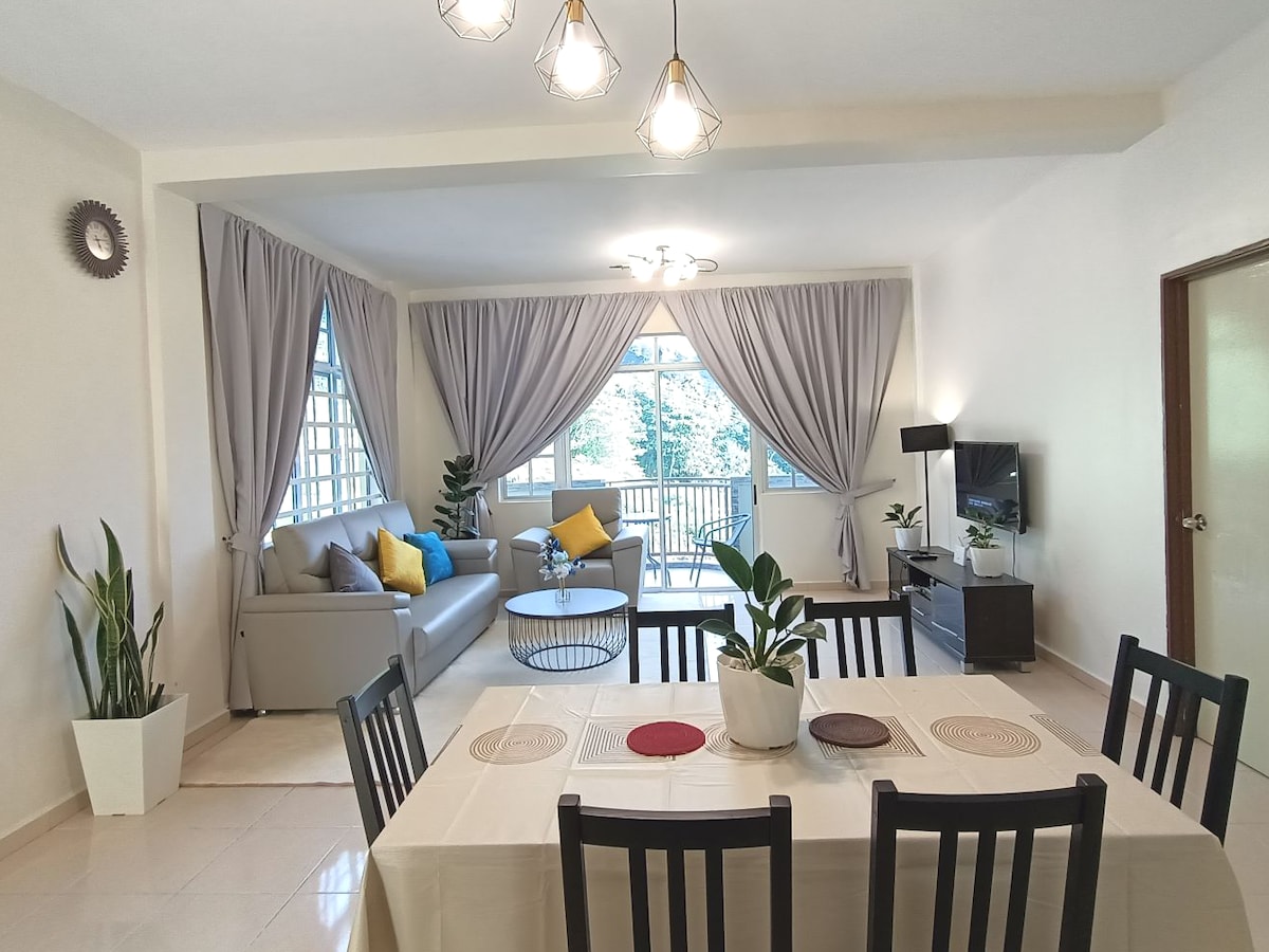 A bright and airy living space features a comfortable gray sofa paired with yellow accent cushions. A glass coffee table sits in front of the sofa, with a small plant adding a natural touch. A dining table with black chairs is placed in the foreground.