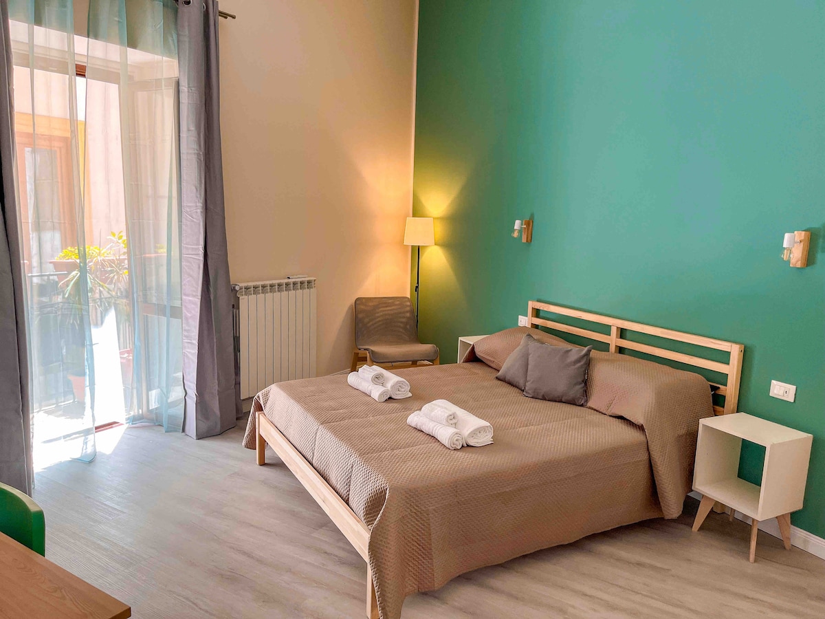 A spacious bedroom features a wooden bed with a neutral-toned quilt, complemented by light grey towels neatly arranged. Large windows allow natural light to fill the room, enhancing the calming green accent wall. A comfortable chair and a lamp provide additional seating and illumination.