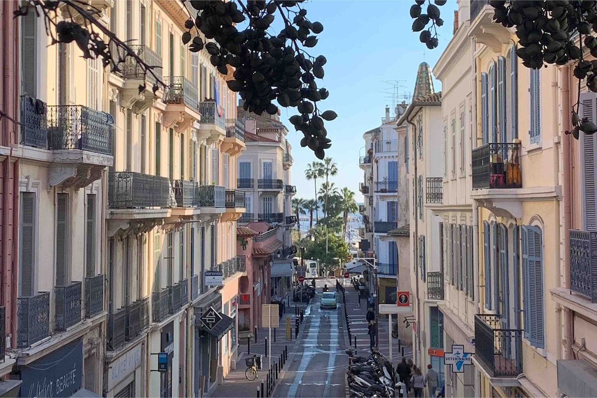 A vibrant street scene in the old town features charming buildings with elegant balconies. The view extends towards the beach, framed by palm trees under a clear sky. Pedestrians and vehicles can be seen navigating the lively pedestrian-friendly area.