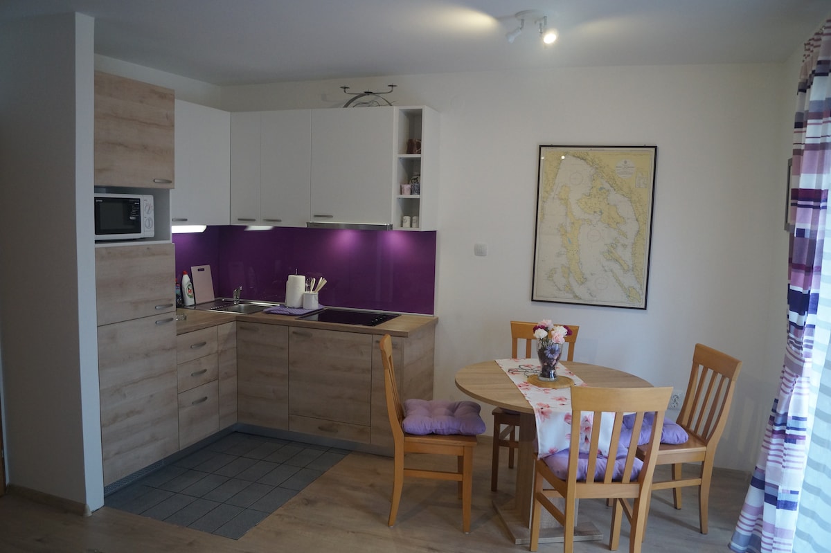 A compact kitchen area features modern cabinetry with light wood finishes. Accents of dark purple paint add a touch of color. A round dining table is set with four wooden chairs, and natural light enters through a window, enhancing the inviting space.