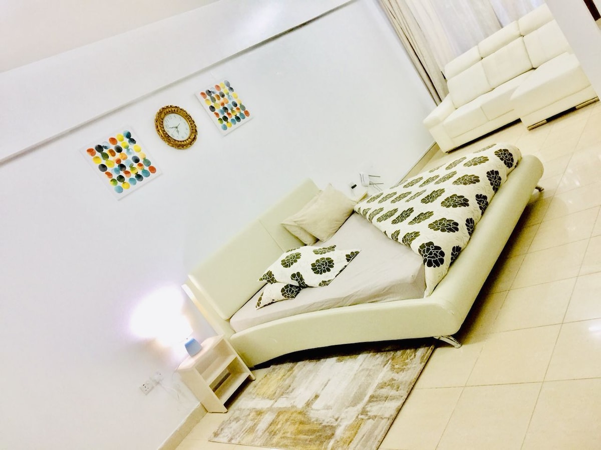 A modern studio space features a queen bed with a floral-patterned bedspread. Colorful wall art adds a touch of vibrancy, and a decorative clock is positioned above the bed. A light-colored sofa and a small bedside table complement the bright interior.