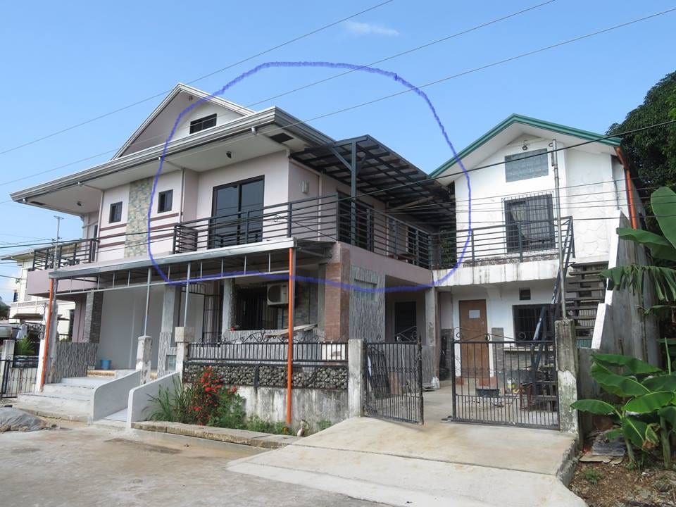 A two-story building with a contemporary design features a spacious balcony with a railing on the upper level. The ground level presents a covered area with a textured stone facade. Surrounding greenery and a well-maintained driveway add to the inviting exterior.