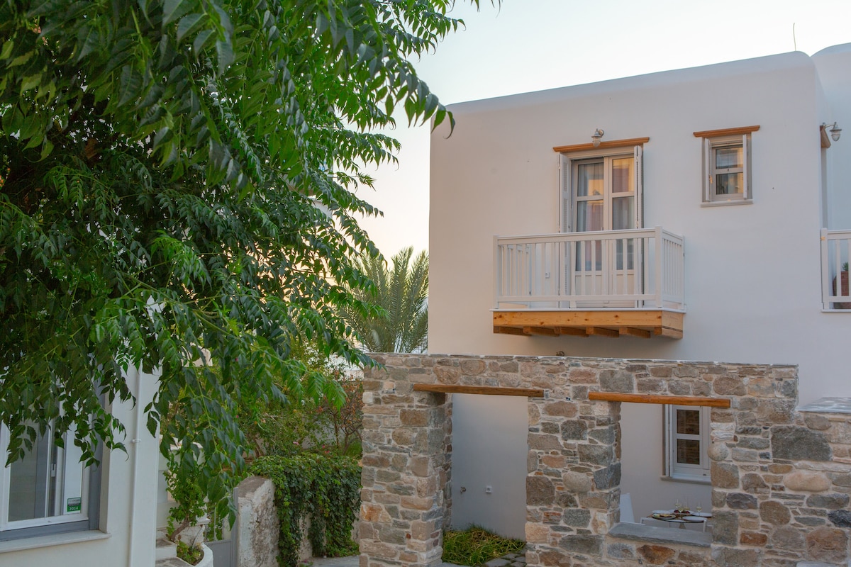 A charming exterior view of the apartment features a light-colored building with large windows and a small balcony above a stone archway. Lush greenery surrounds the structure, creating a serene atmosphere. The stone arch leads towards a pathway, enhancing the inviting entrance.