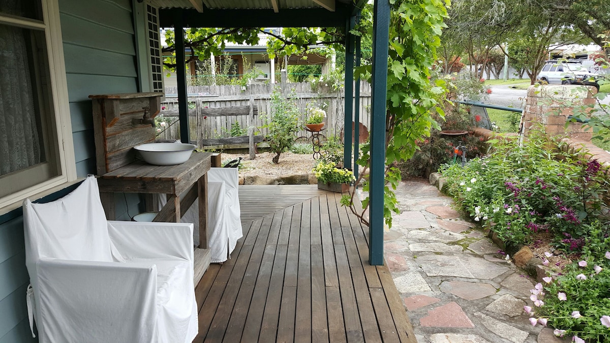 An outdoor area features wooden flooring and is shaded by a roof. White-covered seating is arranged nearby, with a rustic table and wash basin. Lush green vines and a variety of blooming flowers accentuate the garden, creating a serene space for relaxation.