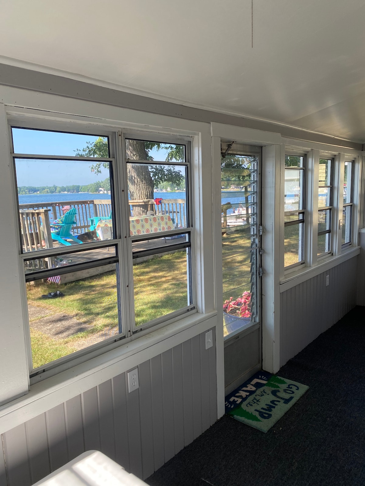 An enclosed porch features large windows providing views of Fish Lake. Natural light fills the space, illuminating the walls and carpet. A door leads to the outdoor deck area, where outdoor seating can be seen. The porch offers a cozy spot for dining or relaxation.