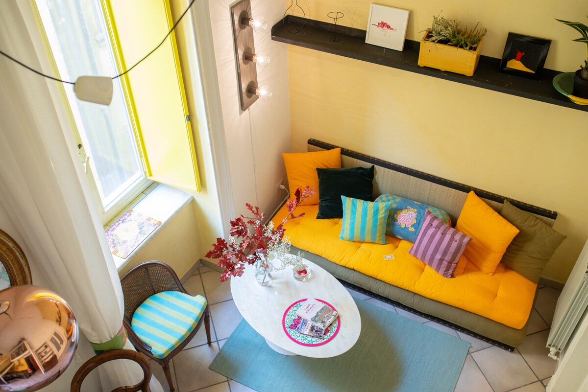 A welcoming living area features a spacious yellow sofa adorned with colorful cushions. A round coffee table sits in front, decorated with a magazine and a small floral arrangement. Bright yellow windows invite natural light, enhancing the cheerful atmosphere of the room.