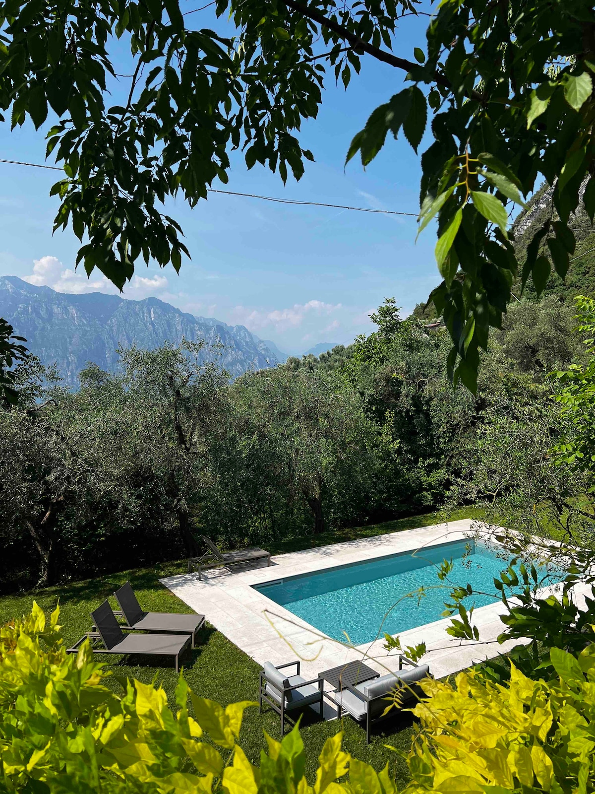A serene outdoor area features a swimming pool surrounded by lush greenery and trees. Sun loungers are positioned nearby, offering a comfortable space to relax. The scenic mountains are visible in the background, enhancing the tranquil setting.