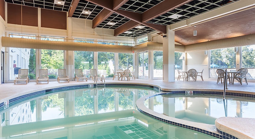 An indoor pool area is highlighted, featuring a curved pool with clear water. Surrounding the pool, several loungers are positioned along the deck, with tables and chairs visible in the background, all under large windows that allow natural light to fill the space.