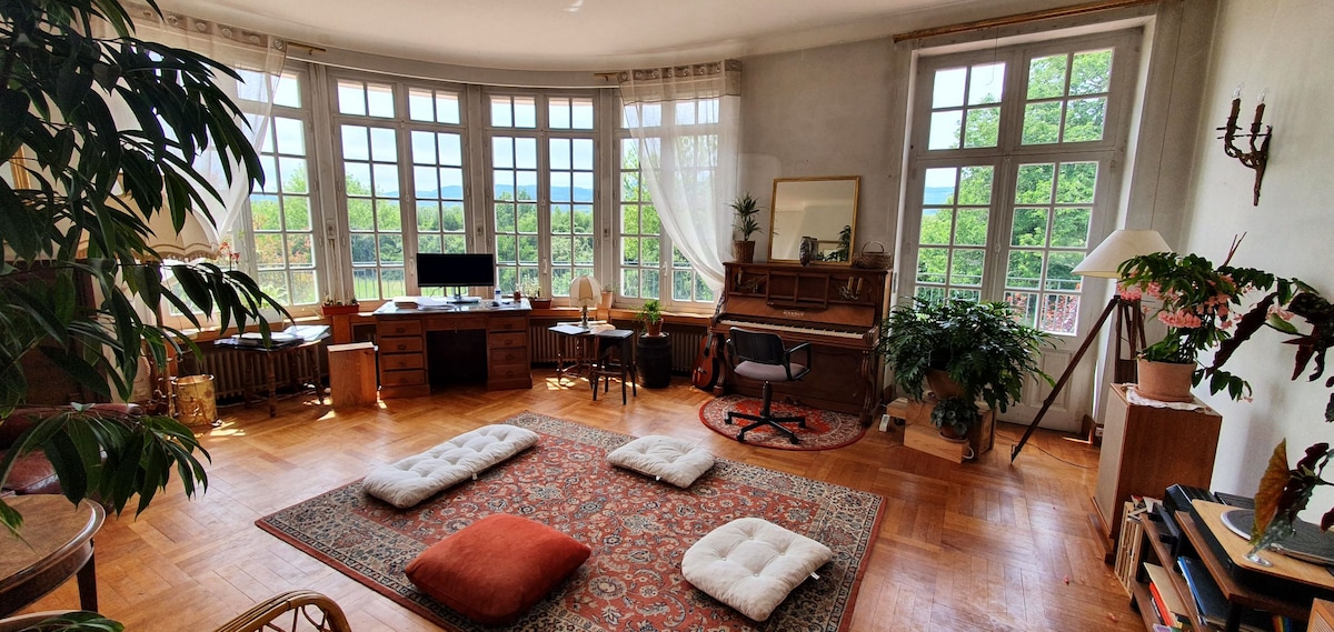 A spacious room features large windows allowing natural light to fill the space. A mix of seating options, including floor cushions and a stylish armchair, is present. A piano and a desk are positioned against the wall, enhancing the inviting aesthetic of the room.