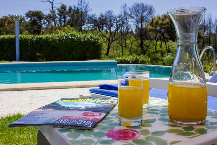 A table set outdoors displays two glasses of orange juice beside a pitcher. A magazine lies open on the table, with a swimming pool and lush greenery visible in the background.