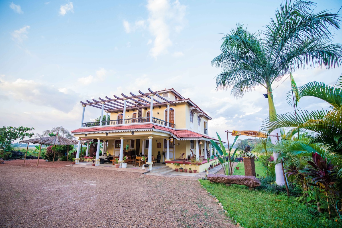 A two-story bungalow with a terracotta roof is surrounded by lush greenery and palm trees. A spacious porch with decorative plants invites relaxation, while the open terrace features pergola structures. The landscape includes a gravel area, enhancing the serene atmosphere of the property.