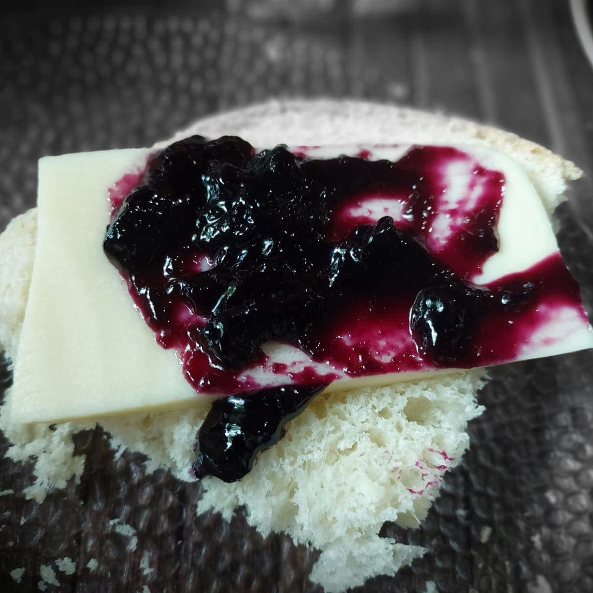 A slice of bread is topped with a layer of cheese and a generous amount of dark berry jam. The soft texture of the bread is visible, contrasting with the glossy sheen of the jam and the smooth surface of the cheese.