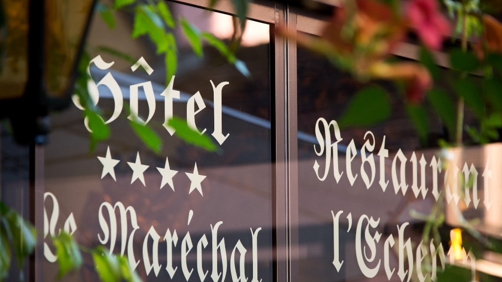 The exterior signage of the hotel is featured prominently, displaying the name 'Hôtel Le Maréchal' along with 'Restaurant l'Echanson.' The lettering is elegantly crafted, partially obscured by lush greenery, suggesting a welcoming and tranquil atmosphere.
