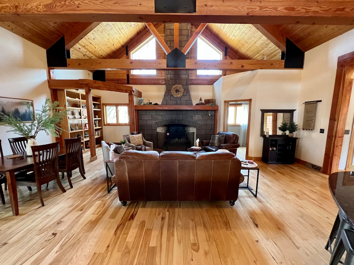 A spacious living area showcases vaulted wooden ceilings and a grand stone hearth. Comfortable seating is arranged around a coffee table, with a dining area visible in the background. Natural light enters through large windows, highlighting the wood flooring and inviting ambiance.