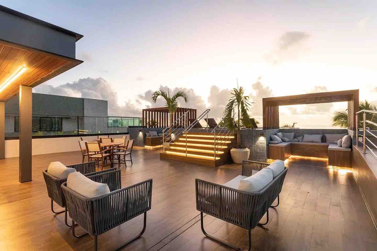 An inviting rooftop terrace features comfortable seating arranged around a wooden deck. Decorative plants enhance the atmosphere. An illuminated staircase leads to an elevated lounging area, surrounded by a scenic view. A dining table is positioned for gatherings, with ample space for relaxation.