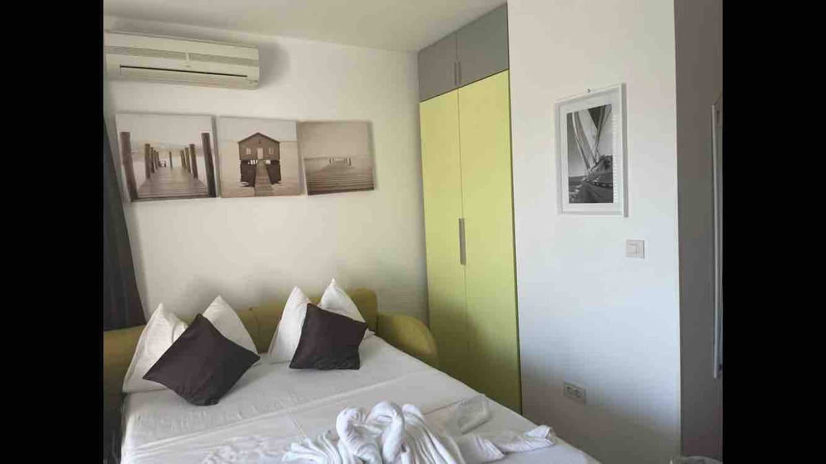 A spacious bed is positioned against a white wall, adorned with two decorative pillows and a comfortable quilt. A closet with a bright yellow finish stands nearby, complemented by black and white photographs on the wall. An air-conditioning unit is mounted above.